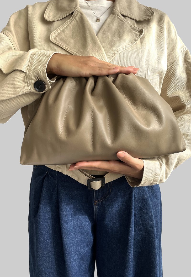 Béžová kožená kabelka s nabraným designem, kterou drží osoba v světle barevné bundě a tmavě modrých džínách. Hladký povrch.