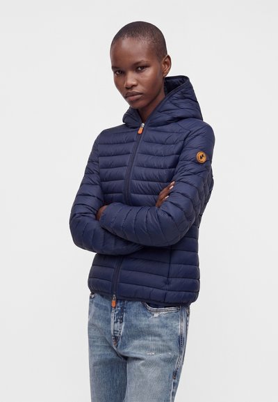 Femme aux cheveux courts portant une veste matelassée à capuche bleu marine et un jean bleu clair, debout les bras croisés devant un fond uni.