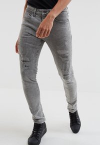 Jean en denim gris slim fit avec des détails déchirés sur les cuisses, affichant un design classique à cinq poches et une fermeture à bouton à la taille.