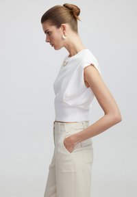 Top recortado blanco con tela drapeada y mangas cortas, combinado con pantalones de cintura alta beige que cuentan con bolsillos laterales y detalles de botones.