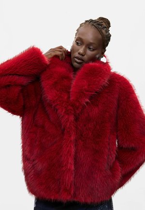 Donna con capelli intrecciati che indossa grandi orecchini a cerchio bianchi e un vivace cappotto in pelliccia ecologica rossa su uno sfondo chiaro e semplice.