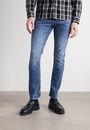 Mann trägt blaue Slim-Fit-Jeans, schwarze Socken, schwarze Leder-Loafers mit metalldetailierter Spange und ein schwarz-weiß kariertes Hemd.
