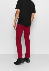 Pantaloni di cotone rossi con una vestibilità slim, abbinati a una camicia nera a maniche corte e sneakers nere, visti da dietro, su uno sfondo bianco.