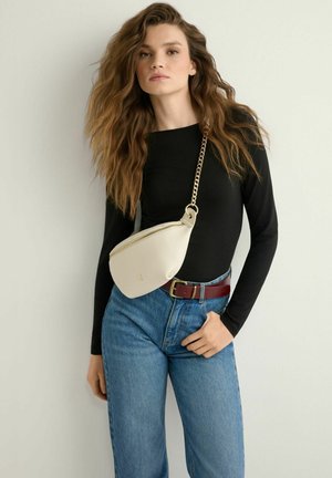 Młoda kobieta o długich, falowanych włosach nosi czarny top, niebieskie dżinsy, brązowy pasek oraz kremową torebkę na ramię z złotym łańcuszkiem.