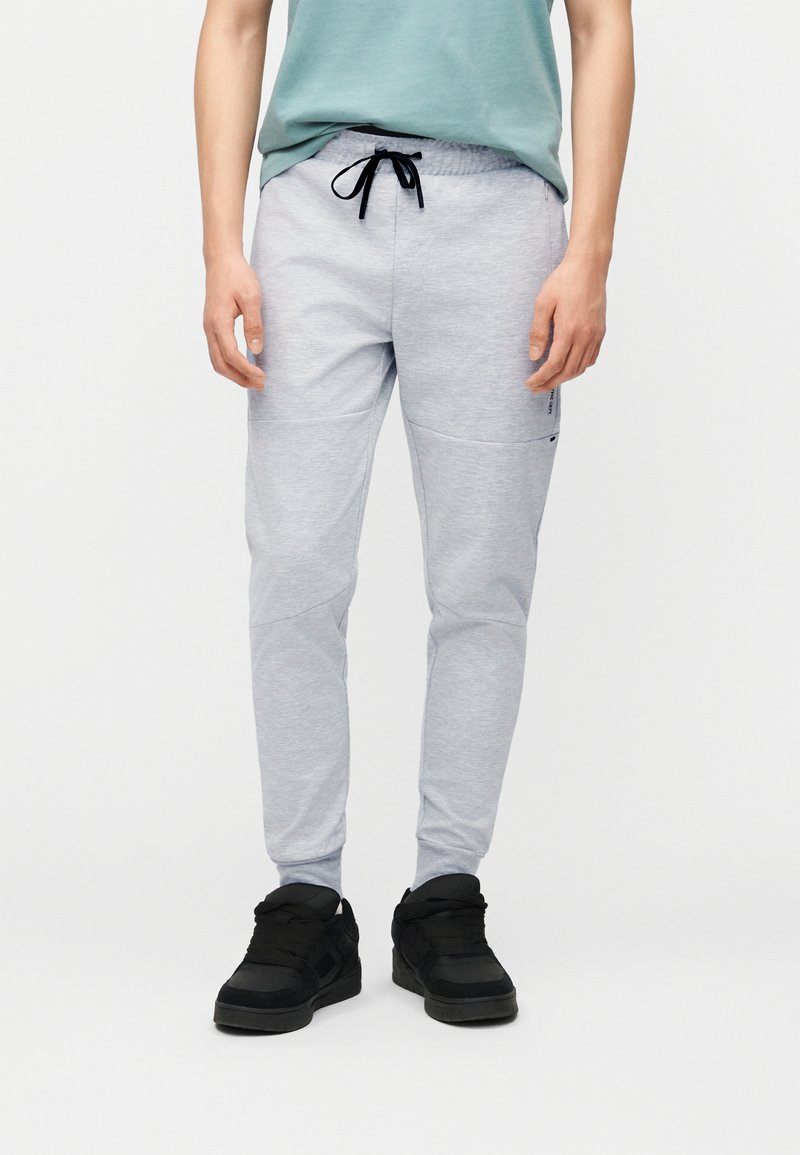 Άτομο που φοράει ανοιχτό γκρι φόρμες με μαύρο κορδόνι και μαύρα αθλητικά παπούτσια, στέκεται μπροστά σε απλό λευκό φόντο.