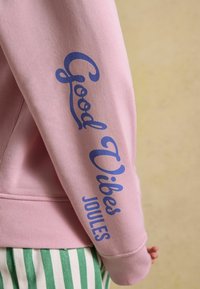 Manche d'un sweat-shirt rose avec texte bleu "Good Vibes" et "Joules", porté avec un pantalon rayé vert et blanc sur fond beige.