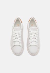 Sneakers bianchi con una finitura in pelle liscia, caratterizzati da lacci bianchi e un accento beige chiaro sul tallone. Design semplice e arrotondato.