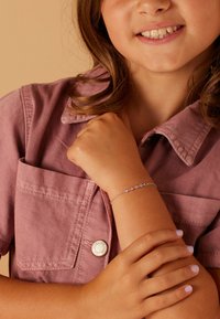 Bracelet features silver chain with pink and purple gems, worn on a child's arm. The shirt is light pink corduroy with a pocket and button.