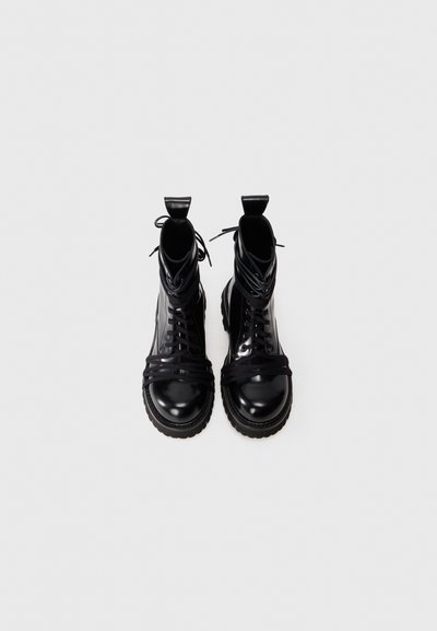 Bottines en cuir noir avec une finition brillante, présentant des orteils ronds, des semelles en caoutchouc épaisses, et des lacets en tissu enroulés autour de la tige.