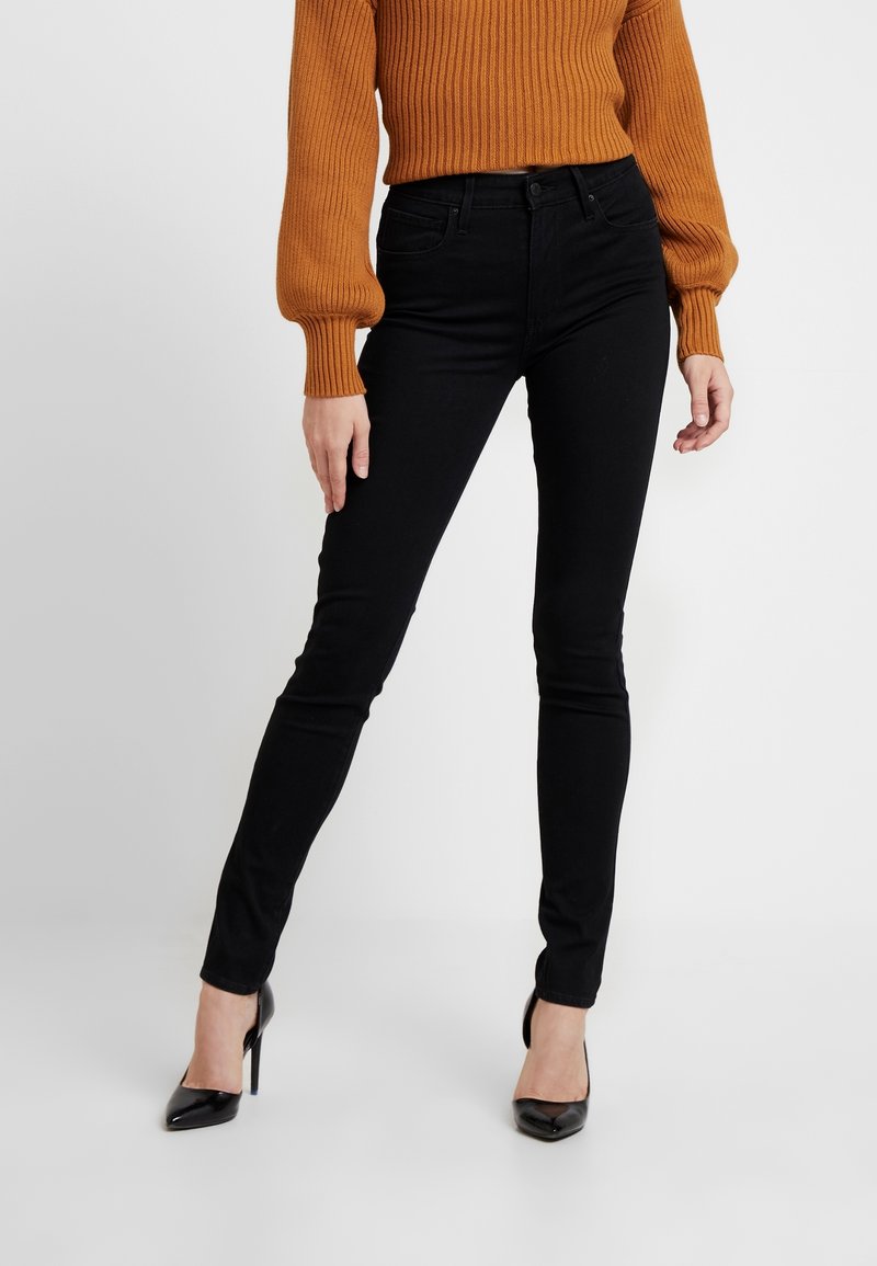 Jean skinny noir à texture lisse, taille haute et coupe ajustée, associé à un pull orange côtelé et des chaussures noires à talons hauts.