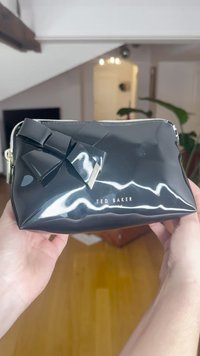 Bolsa de cosméticos preta brilhante com detalhe de laço, fecho prateado e logótipo "TED BAKER" na frente. Textura suave com forma estruturada.