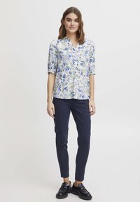 Blouse florale blanche avec des motifs bleus et verts, à manches courtes et col en V, associée à un pantalon slim bleu marine et des chaussures noires.