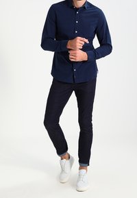 Hombre ajustando el puño de una camisa de cuadros azul oscura, vistiendo jeans oscuros ajustados y zapatillas blancas, de pie contra un fondo blanco.