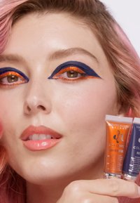 Vloeibare make-up tubes in oranje en blauw gehouden naast een gezicht met roze haar. Dikke eyeliner met marineblauwe en oranje accenten. Glanzende nude lippen.