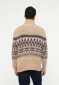 Pull en tricot beige avec un col montant, présentant un motif géométrique bleu sur la poitrine et des manches évasées, affichant une finition texturée.