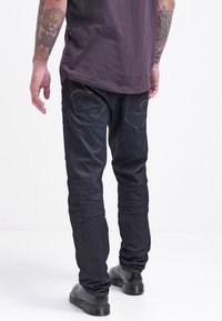 Vaqueros de mezclilla oscuro con un ajuste slim, que cuentan con cinco bolsillos y detalles de costura en color claro. Combinados con una camisa púrpura lisa y botas negras.