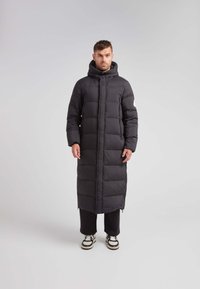 Lang zwart pufferjack met capuchon, voorzien van een knoopsluiting, gestikte ontwerp, zijzakken en een rechte snit. Draag het over donkere broeken en sneakers.