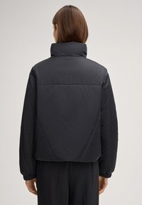 Schwarze gepolsterte Jacke mit hohem Kragen, tailliertem Design, elastischen Bündchen und verdeckten Nahtdetails in einer glatten, matten Textur.