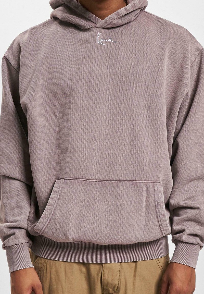 Mężczyzna w zakurzonej fioletowej bluzie z kapturem z przednią kieszenią i małym białym logo, w połączeniu z jasnobrązowymi spodniami, widoczny tułów, bez widocznej twarzy.