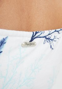 Tissu blanc avec un motif de corail bleu clair et bleu foncé, comportant une petite étiquette translucide portant "Pepe Jeans" près de la peau.