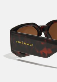 Occhiali da sole tartarugati con lenti marroni, caratterizzati da un design geometrico spesso. Nome del marchio giallo "PRIVÉ REVAUX" stampato sulla stanghetta.