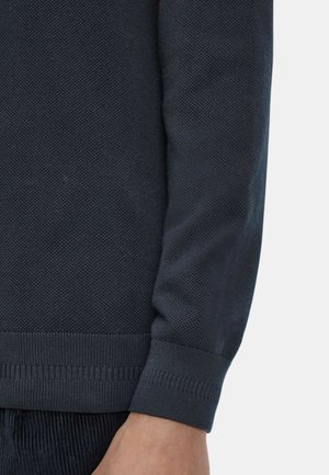 Dunkelblauer, strukturierter Strickpulloverärmelsaum mit geripptem Bündchen am Arm einer Person vor hellem Hintergrund.