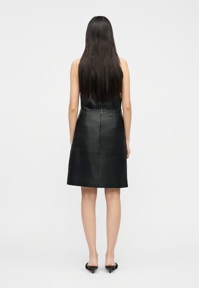 Γυναίκα με μακριά σκούρα μαλλιά που φοράει ένα μαύρο δερμάτινο φόρεμα χωρίς μανίκια και μαύρα mule με τακούνι, όρθια με την πλάτη προς το μέρος μας σε απλό φόντο.