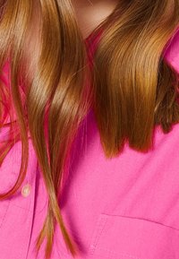 Roze shirtstof met een zachte textuur, voorzien van een zak aan de rechterzijde. Lange, rechte bruine haren zijn gedeeltelijk zichtbaar langs de randen.