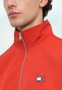 Κόκκινο μπουφάν με φερμουάρ και ψηλό λαιμό, φτιαγμένο από λείο ύφασμα. Διαθέτει ένα μικρό, πολύχρωμο λογότυπο "TOMMY JEANS". Λεπτομέρεια από ασημί φερμουάρ.