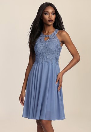 Frau in einem ärmellosen, knielangen blauen Kleid mit Spitzenoberteil und fließendem Rock, steht mit der Hand an der Hüfte vor einem neutralen Hintergrund.