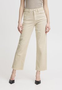 Jeans beige a vita alta con design a gamba dritta, realizzati in denim; presentano tasche frontali e dettagli in metallo argentato, abbinati a tacchi neri a punta.