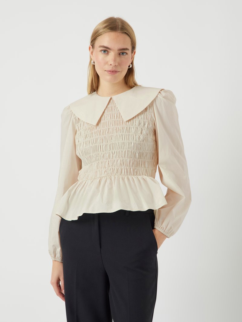 Cremefarbene Bluse mit großem Kragen, gesmoktem Oberteil und gerafftem Peplum-Saum. Lange Ärmel mit elastischen Bündchen. Tragen Sie sie mit schwarzen Hosen.
