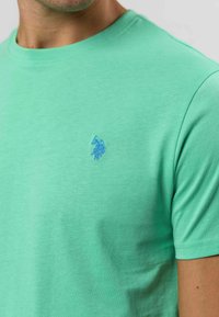 Müntroheline lühikeste varrukatega T-särk, mis on valmistatud pehmest puuvillast, väikese sinise tikandiga logo vasakul rinnal ja klassikalise ringkaelusega.