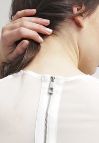 Naisen käsi vetää takaisin ruskeita hiuksia, paljastaen niskan ja hopeisen vetoketjun valkoisen läpikuultavan paidan takana.
