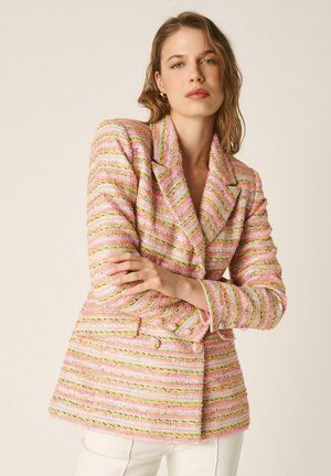 Mujer con cabello castaño claro que lleva una blazer texturizada de rayas en tonos pastel rosa, con los brazos cruzados, de pie frente a un fondo beige liso.