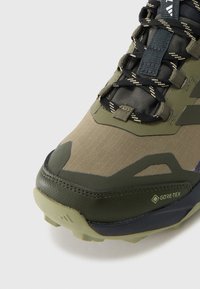 Zöld és barna szintetikus felsőrésszel készült kültéri cipő, texturált dizájnnal, megerősített orrésszel és GORE-TEX márkajelzéssel a vízállóság érdekében.