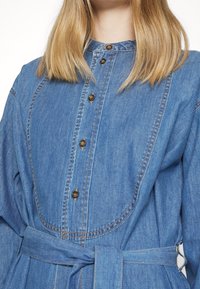 Denim dress featuring a button-down front, a cinched waist with a fabric belt, and a round neckline. Light blue color with textured fabric.