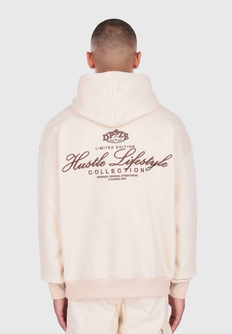 Homme portant un sweat à capuche crème avec la capuche sur la tête, texte "Collection Mode Travailleur" et d'autres marques imprimées au dos en lettres brunes.