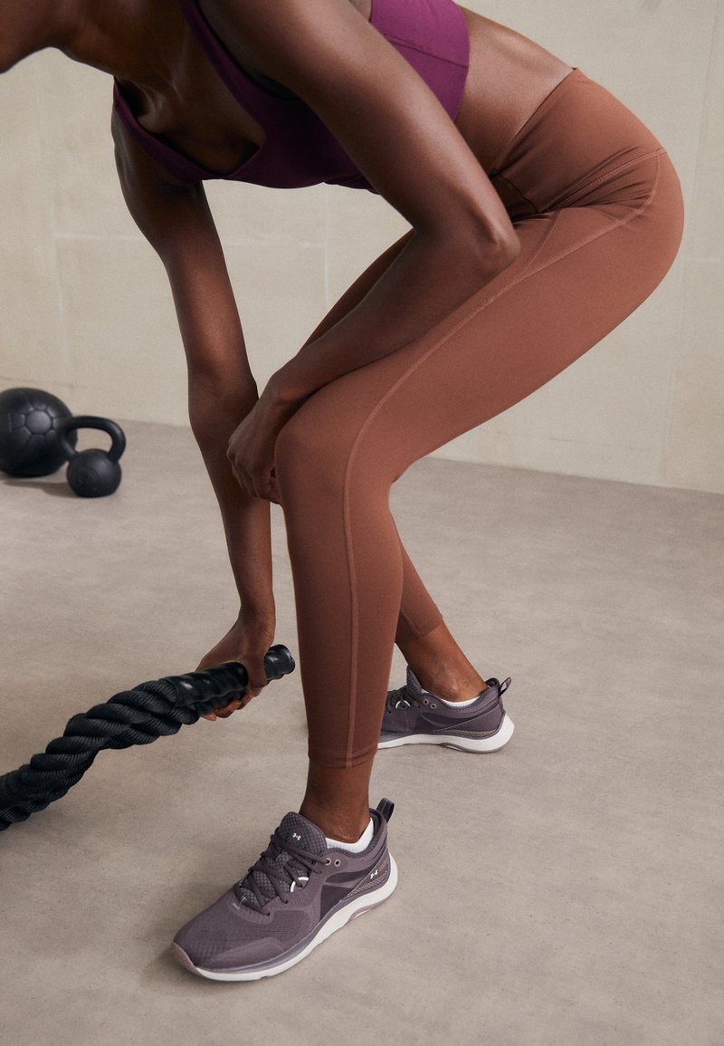 Brūnas sportiskās leggingas, purpura sporta krūšturis un pelēki sporta apavi. Gluda auduma, pieguļošs dizains un teksturēta saķere uz virves.