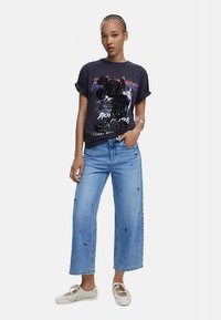 Fekete grafikás póló élénk mintával és laza ujjakkal; világos koptatott, bő szárú farmer hímzett részletekkel; fehér tornacipő.