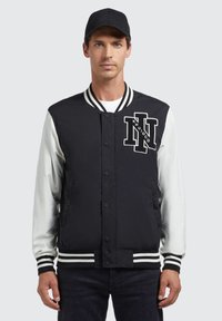 Mustvalge varsity-jakk, millel on triibuline soonik ja ääreosa, esiküljel tikandiga logo ja vastandlikud valged varrukad.
