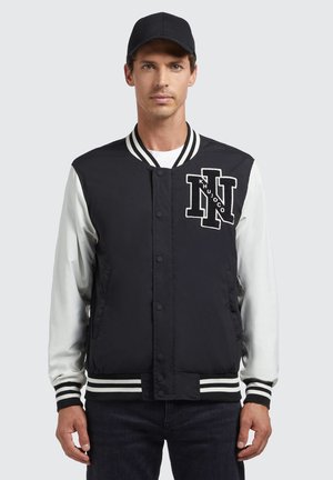Črno-bela varsity jakna z črtastimi rebrastimi manšeti in robom, ki ima izšito veliko na prsih ter kontrastne bele rokave.