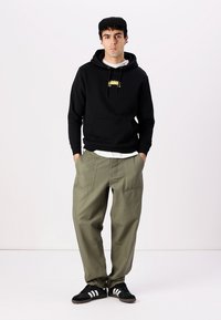 Black hooded sweatshirt with a gold logo, layered over a white shirt, paired with loose olive green cargo pants and black sneakers.