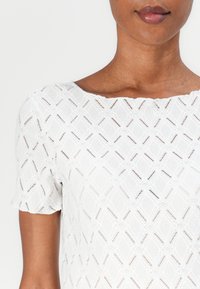 Blusa bianca con texture che presenta un motivo a rombi, maniche corte e scollo a barca. Il tessuto ha una qualità trasparente e dettagli delicati.