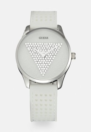 Montre-bracelet Guess blanche avec lunette argentée, triangle texturé de points sur le cadran, aiguilles blanche des heures, minutes et secondes, et un bracelet blanc perforé.