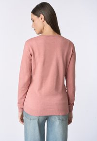 Maglione a maglia rosa chiaro con maniche lunghe, scollo rotondo e orlo a coste. Realizzato in un materiale morbido, indossato sopra jeans azzurri chiari.