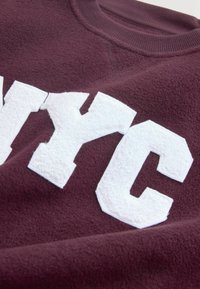 Maroon fleece-sweatshirt, jossa on suuri, teksturoitu valkoinen "NYC" -kirjoitus, pyöreä kaula-aukko ja pehmeä, harjattu kangas.