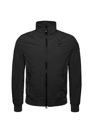 Chaqueta negra con cremallera y cuello alto, con bolsillos laterales, puños elásticos y un tejido negro texturizado. Presenta un pequeño logotipo amarillo en el pecho.