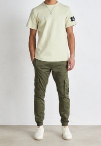 Light yellow short-sleeve shirt with logo patch, olive green cargo pants with side pockets, and white sneakers with eyelets.