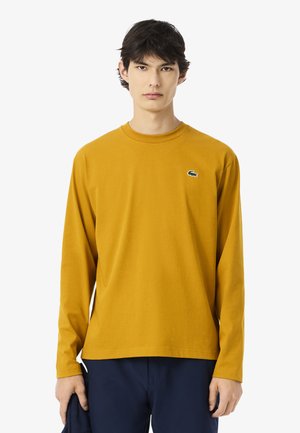 Langarm-T-Shirt in Senfgelb aus Baumwolle, mit rundem Halsausschnitt und kleinem grünen gesticktem Logo auf der Brust.